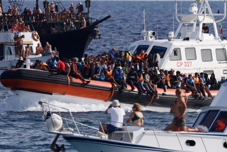 An Italian Coast Guard boat carries migrants as tourists on boat, foreground, watch, near the port of the Sicilian island of Lampedusa, southern Italy, Monday, Sept. 18, 2023. (Cecilia Fabiano/LaPresse via AP)